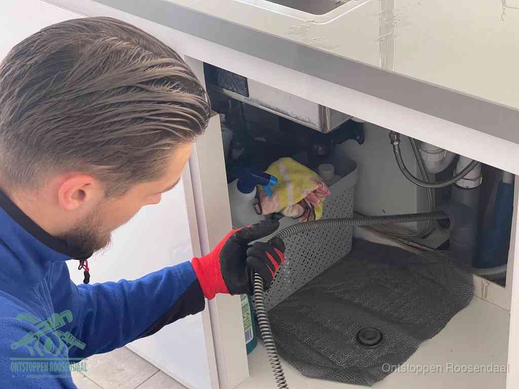 Een monteur voert een flexibele ontstoppingsveer in onder het keukenkastje, langs de sifon, om de gootsteen te ontstoppen in Roosendaal