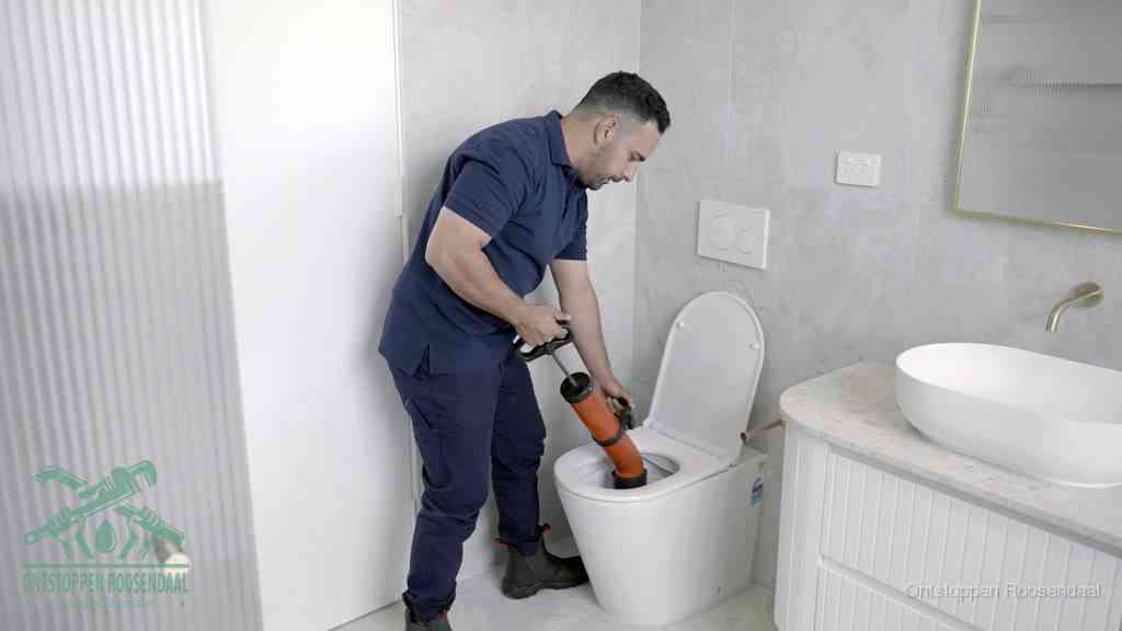 Een monteur buigt over een wandcloset en duwt met de hand een persluchtontstopper in de toiletpot om de verstopping handmatig los te maken in Roosendaal