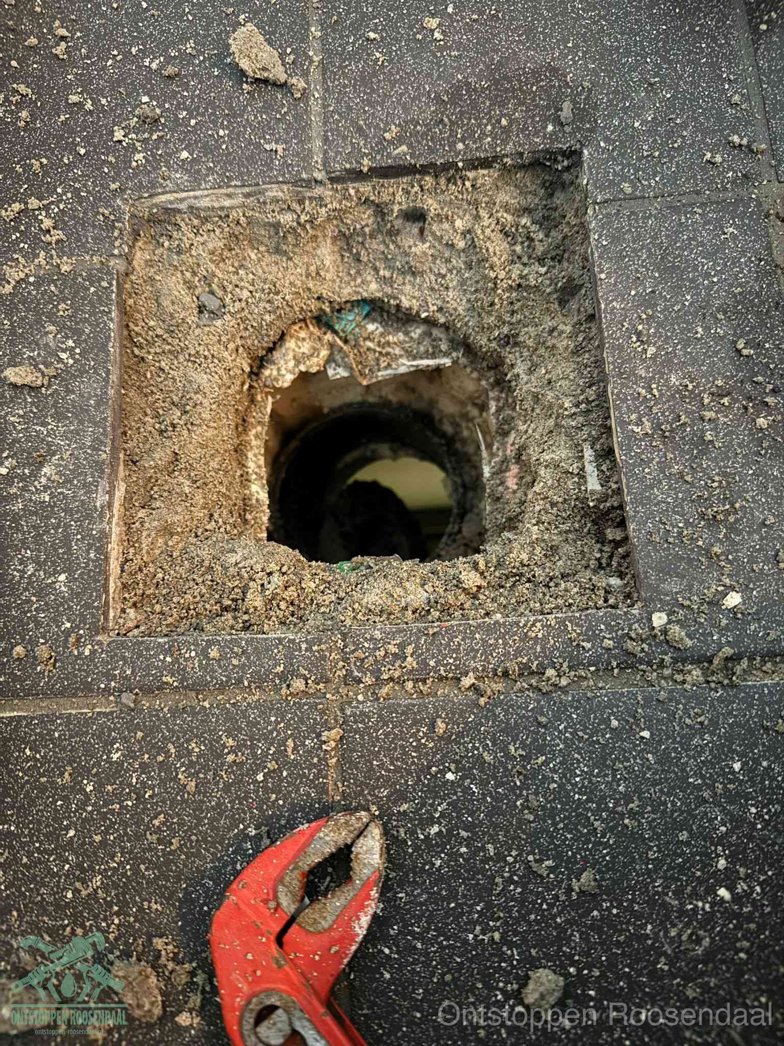 Een geopend rioolputje in een betontegelvloer met losgemaakt rooster en een zichtbare ronde afvoerbuis, het achterhalen waar de afvoerverstopping zit in Roosendaal