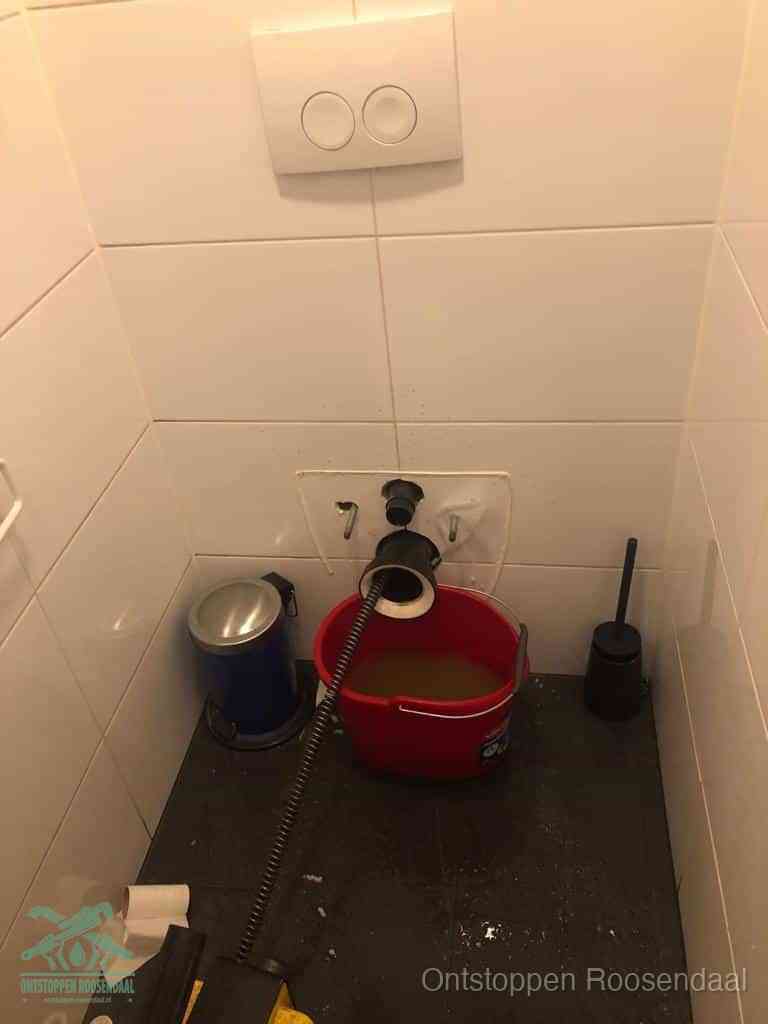 Een ontstopte wand-wc met verwijderde toiletpot waar een ontstoppingsveer in de wanduitgang verdwijnt, het achterhalen waar de verstopping zit in Roosendaal