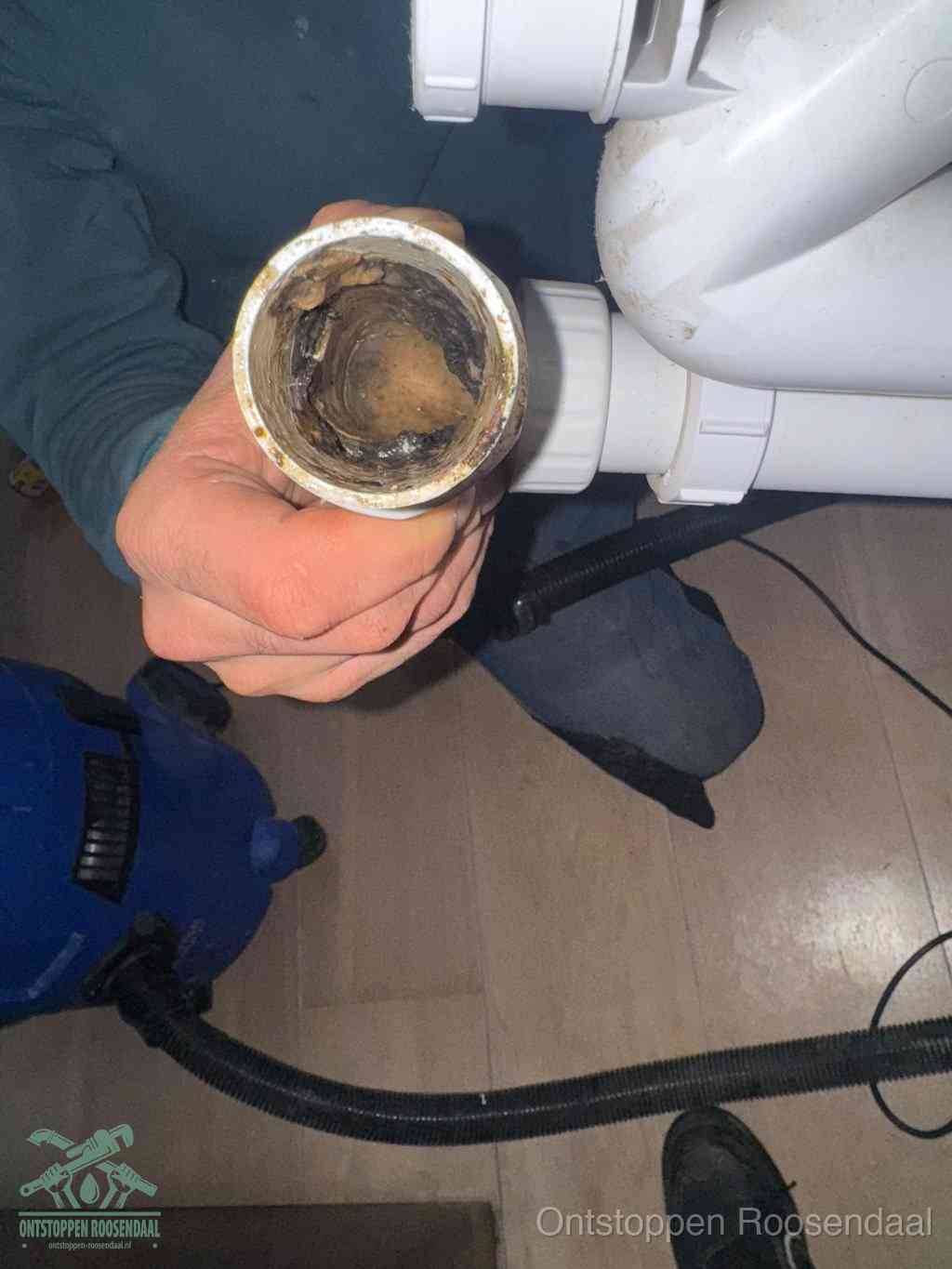 Een hand houdt een losgekoppeld PVC-afvoerbuisstuk vast dat van binnen volledig is dichtgeslibd met vet, vuil en aanslag, de meest voorkomende oorzaak van een verstopte afvoer in Roosendaal