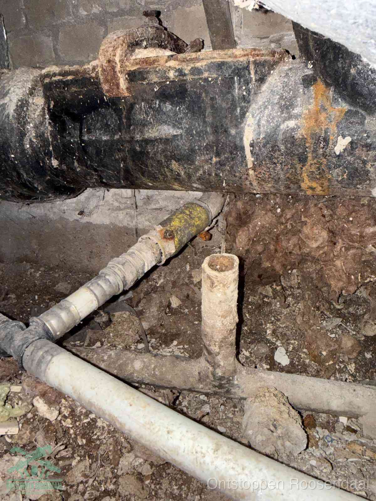 Een dikke zwarte hoofdrioolbuis met roest- en kalkaanslag in een kruipruimte, oorzaak van terugkerende structurele afvoerproblemen in Roosendaal