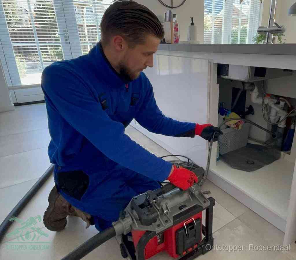 Een monteur met rode handschoenen knielt in een keuken en voert een elektrische ontstoppingsveer in tijdens het professioneel afvoer ontstoppen in Roosendaal