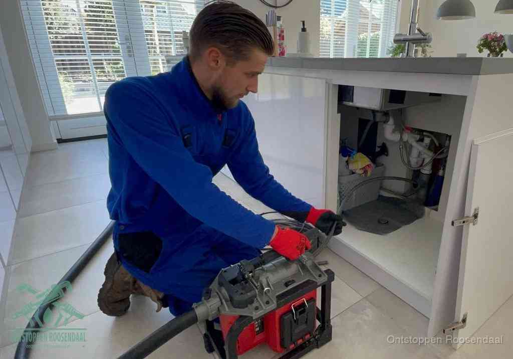 Een monteur voert een professionele elektrische ontstoppingsmachine in de afvoer onder de spoelbak tijdens het gootsteen ontstoppen in Roosendaal